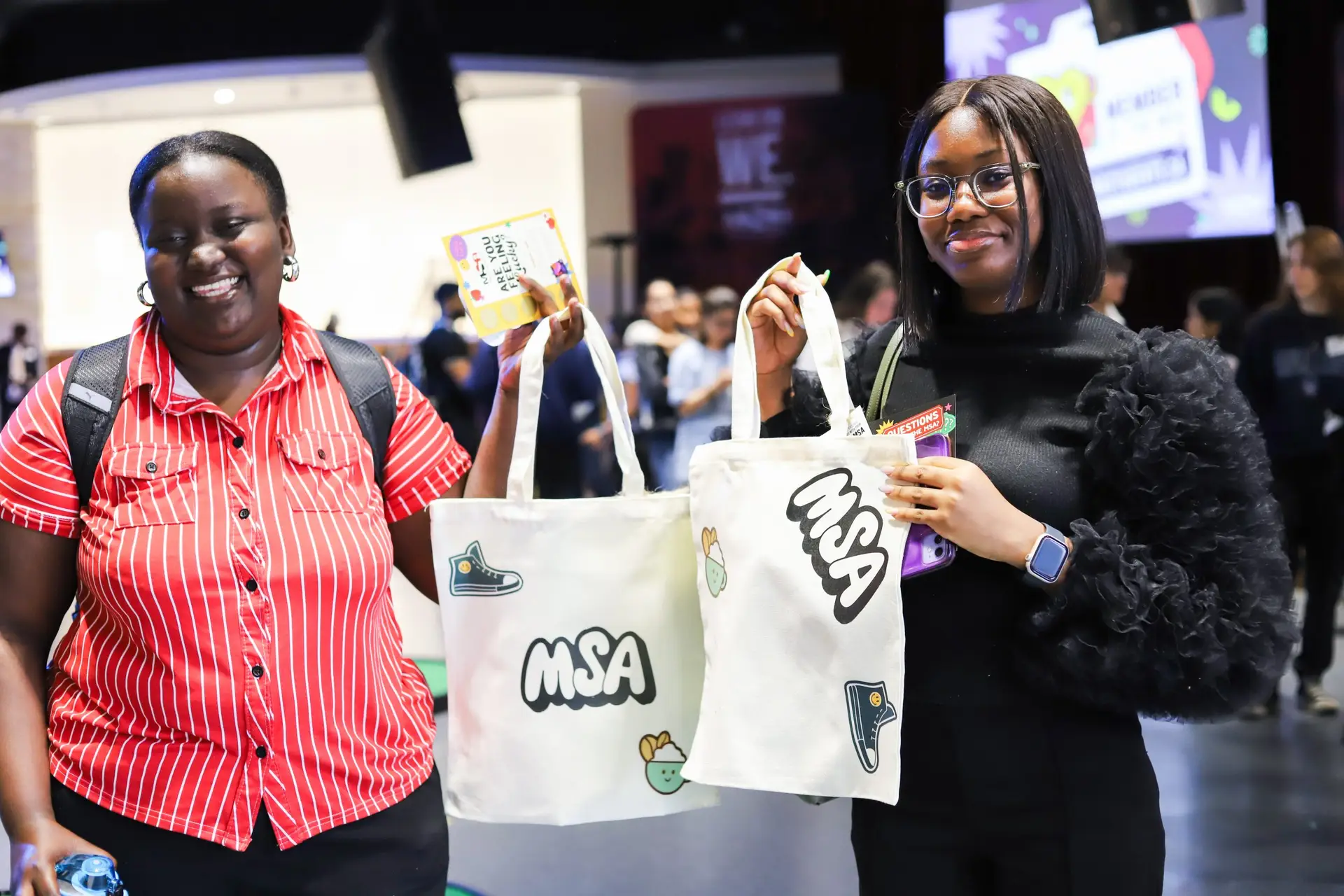 Two smiling students holding MSA Book Bags at a campus event, with other attendees visible in the background.